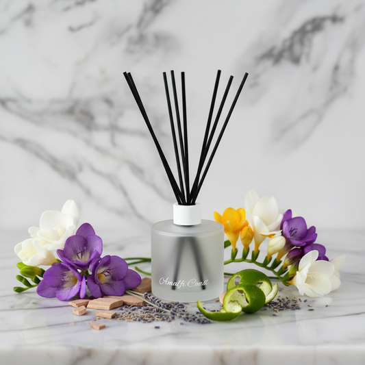 Reed diffuser with black sticks in a clear bottle on a marble surface with purple flowers.