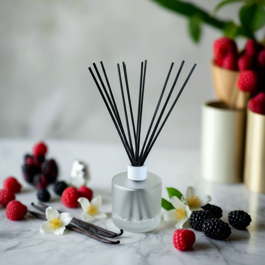 A frosted glass reed diffuser bottle with black reeds stands on a marble surface surrounded by raspberries, blackberries, vanilla beans, and small white flowers. The diffuser label reads 'Black Raspberry'.