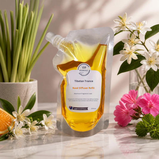 A stand-up refill pouch of Wessentials 'Tibetan Trance' Reed Diffuser Refill, filled with golden-yellow liquid, styled on a reflective surface with green reeds, white flowers, and pink blossoms.