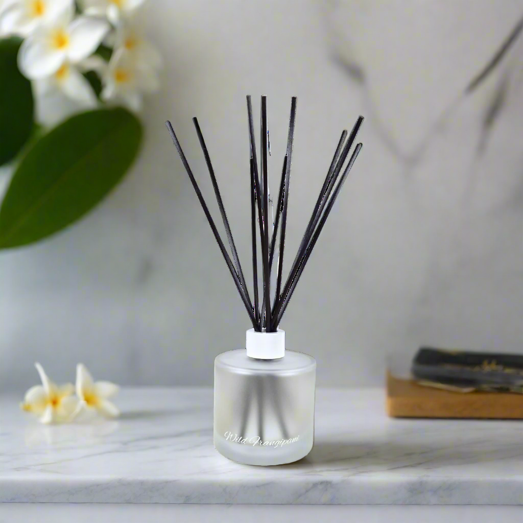 Reed diffuser with black sticks in a clear bottle on a marble surface with flowers in the background