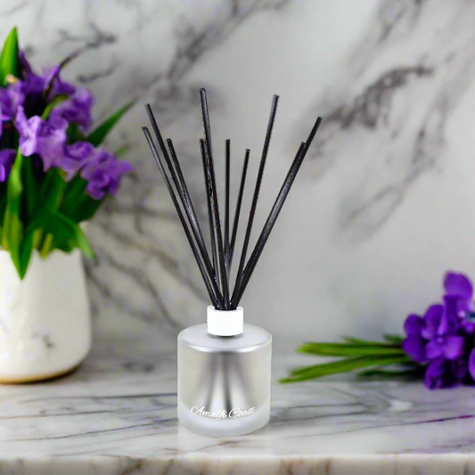 Reed diffuser with black sticks in a clear bottle on a marble surface with purple flowers.