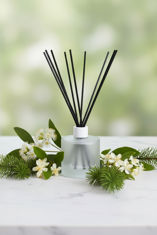 A frosted glass reed diffuser bottle with black reed sticks, styled on a white marble surface with green leaves, pine needles, and small white flowers against a soft green bokeh background.