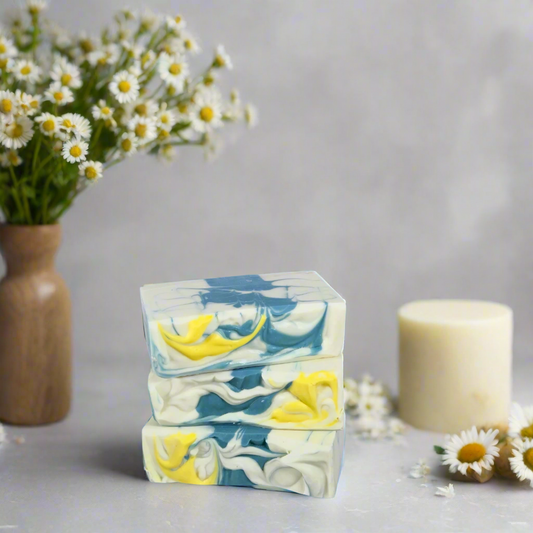 Stack of floral soap bars with a candle and flowers in the background on a light surface.