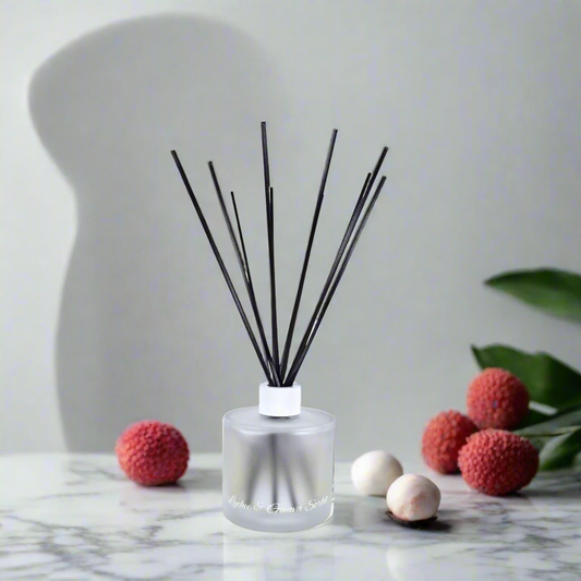Reed diffuser with black sticks in a clear bottle on a marble surface with lychees and leaves.