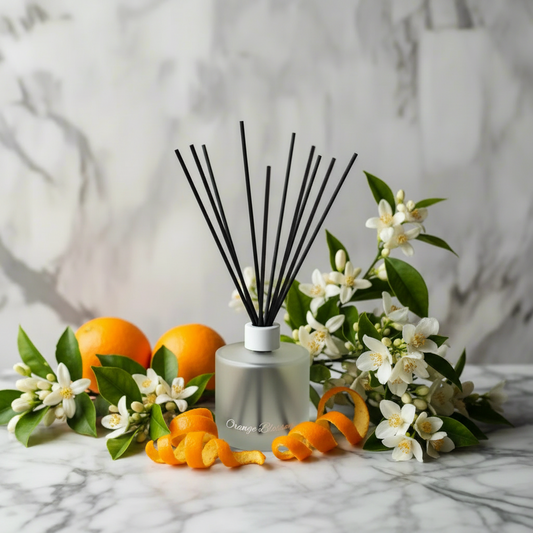 A reed diffuser bottle with black reeds stands on a white marble surface, surrounded by oranges, white blossoms, and orange peel. The diffuser bottle has a brushed silver finish with gold script text near the base.