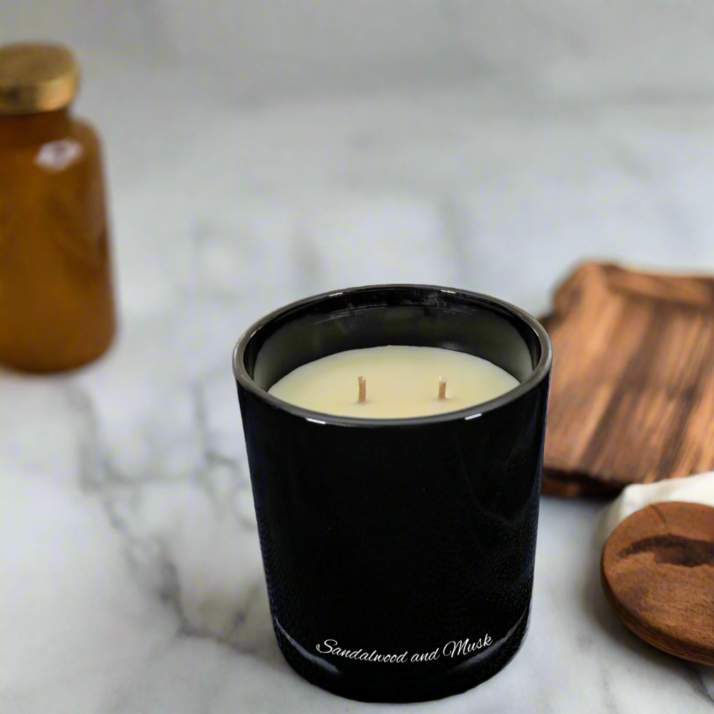 Black candle with sandalwood and musk scent on a marble surface with wooden coaster and bottle.