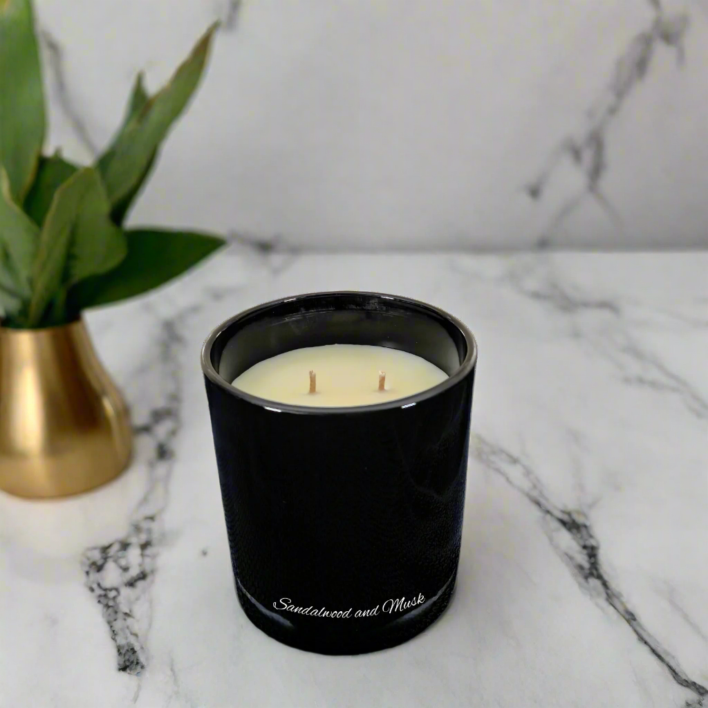 Black candle with 'Sandalwood and Musk' scent on a marble surface next to a gold vase with green leaves.