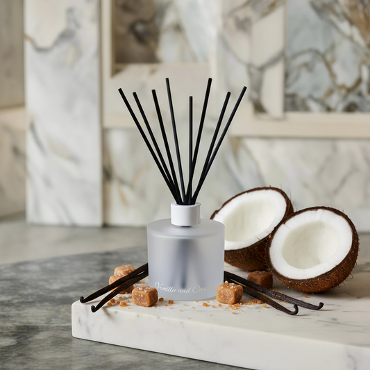 A reed diffuser bottle with black reeds stands on a white marble slab, styled with coconut halves, brown sugar cubes, and vanilla beans against a softly blurred marble background.